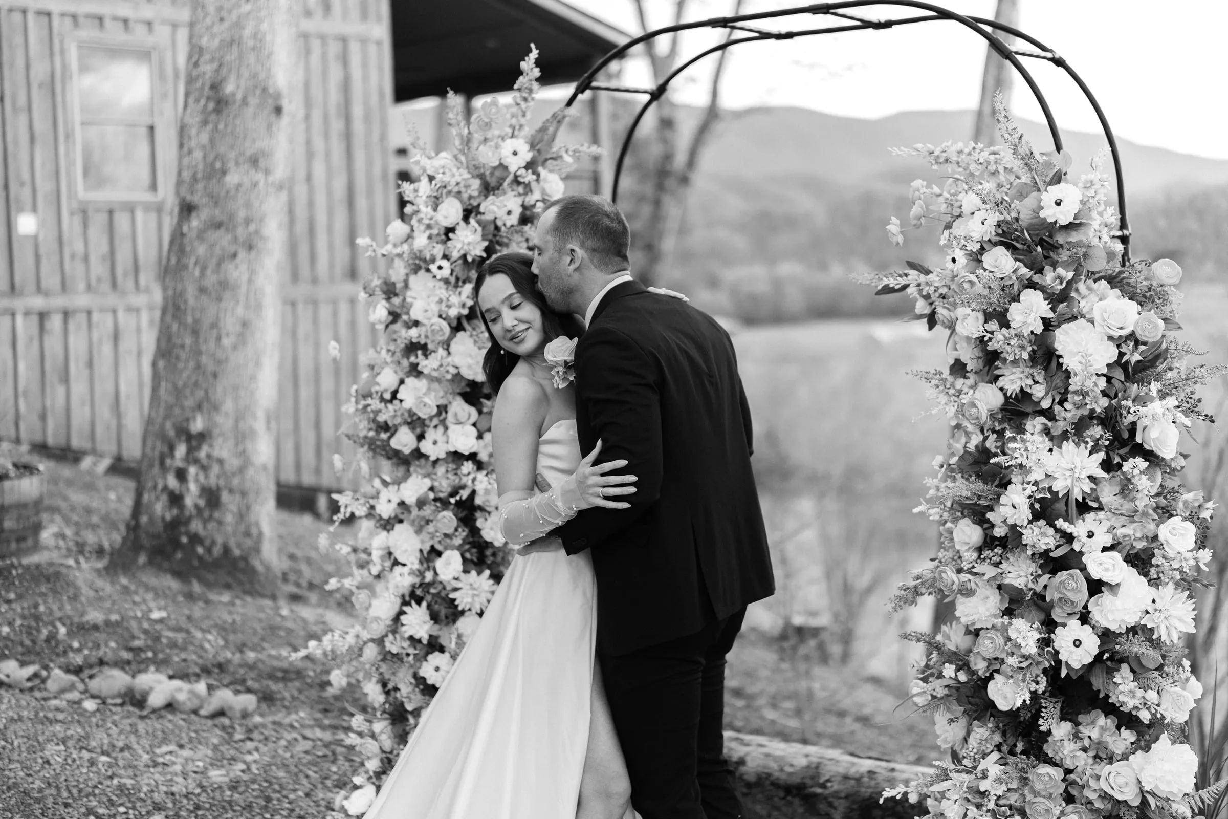 Black-and-white photo of a couple embracing at the floral arch with mountains behind