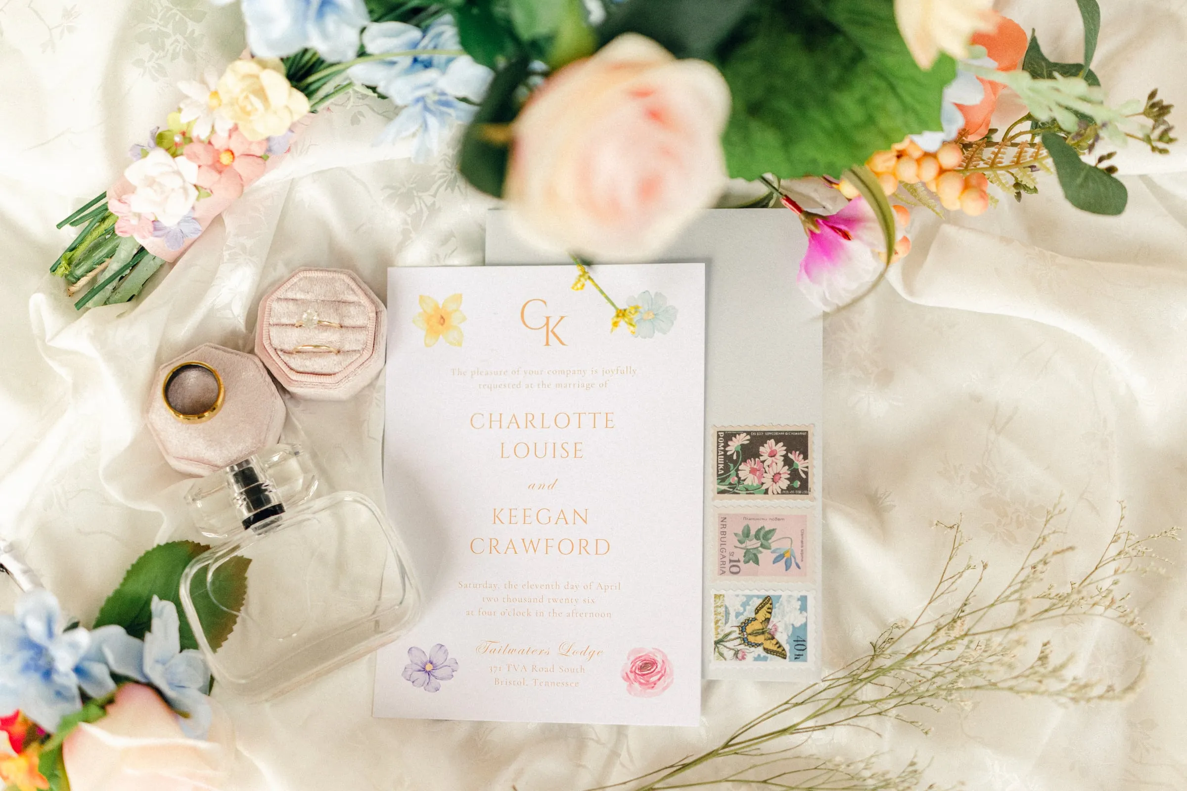 Flat-lay of a monogrammed invitation suite with perfume, stamps, and florals