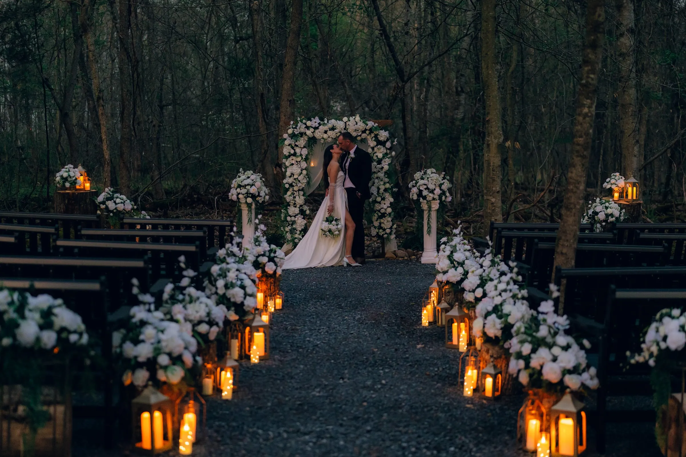Lantern-lined ceremony aisle leading to a floral arch at dusk in the forest at Tailwaters Lodge