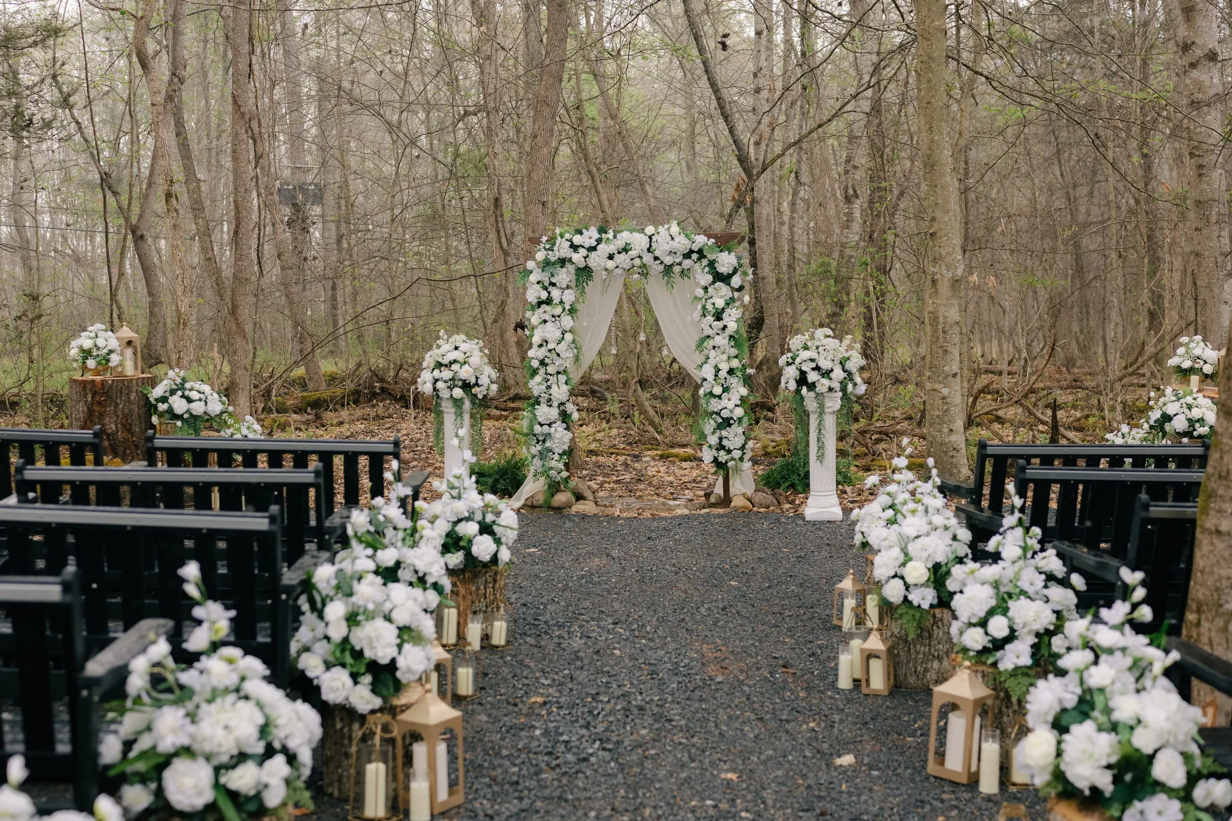Fernwood ceremony arbor with white florals and a lantern-lined aisle in the woods